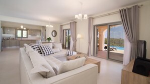 A modern living room with a sofa, coffee table, and a TV stand. There is a kitchen in the background with a window and a dining area. - Villa Aglaia in Mavrata (Kefalonia)