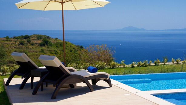 A poolside area with two lounge chairs, a large umbrella, and a view of the sea. - Villa Aglaia in Mavrata (Kefalonia)