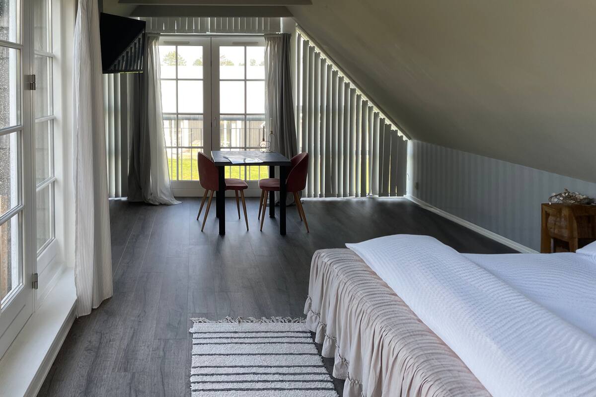 Un dormitorio con una cama grande, una mesa de comedor con sillas y vistas al exterior a través de una ventana con persianas.