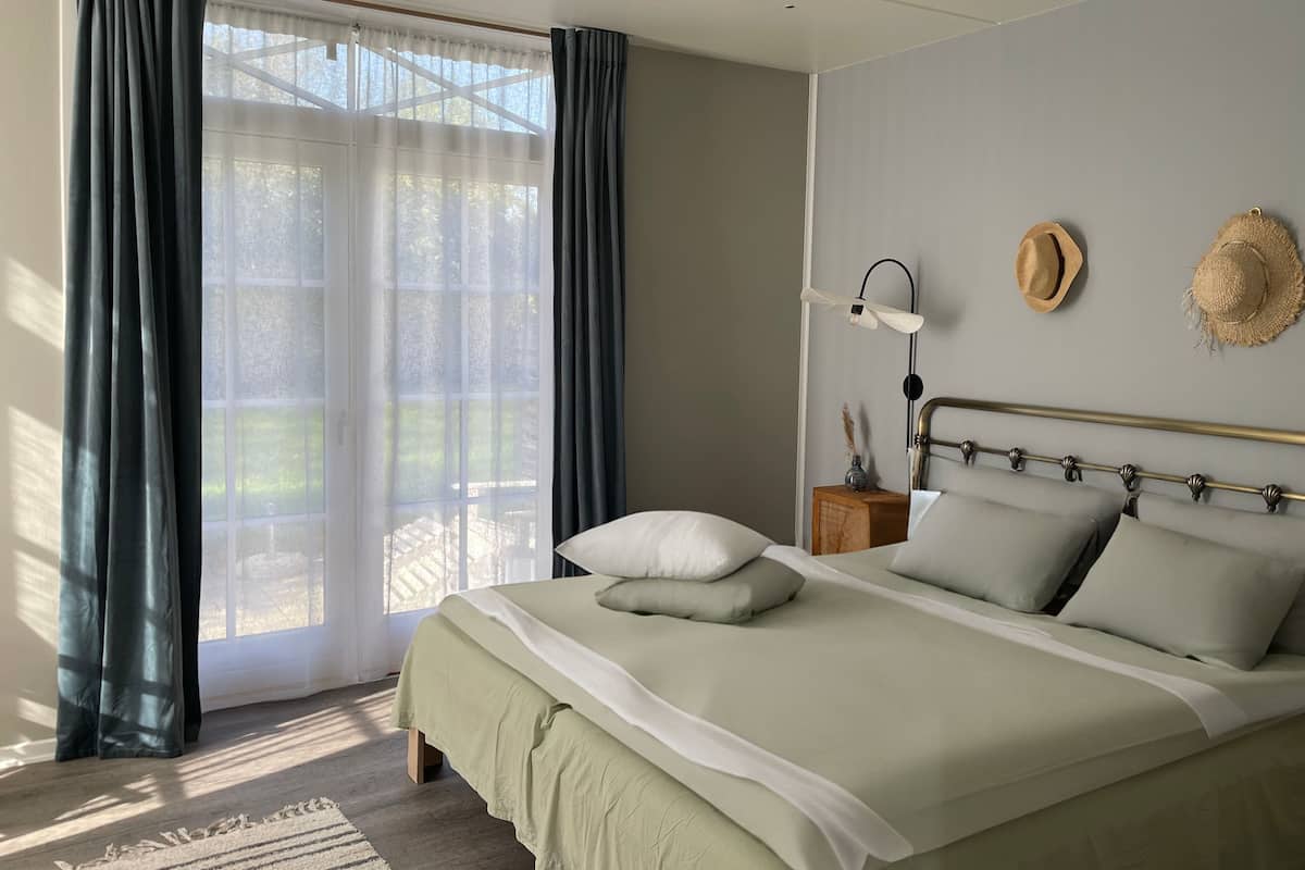 Un dormitorio con cama, cortinas, una mesita de noche, una lámpara y dos sombreros en la pared.