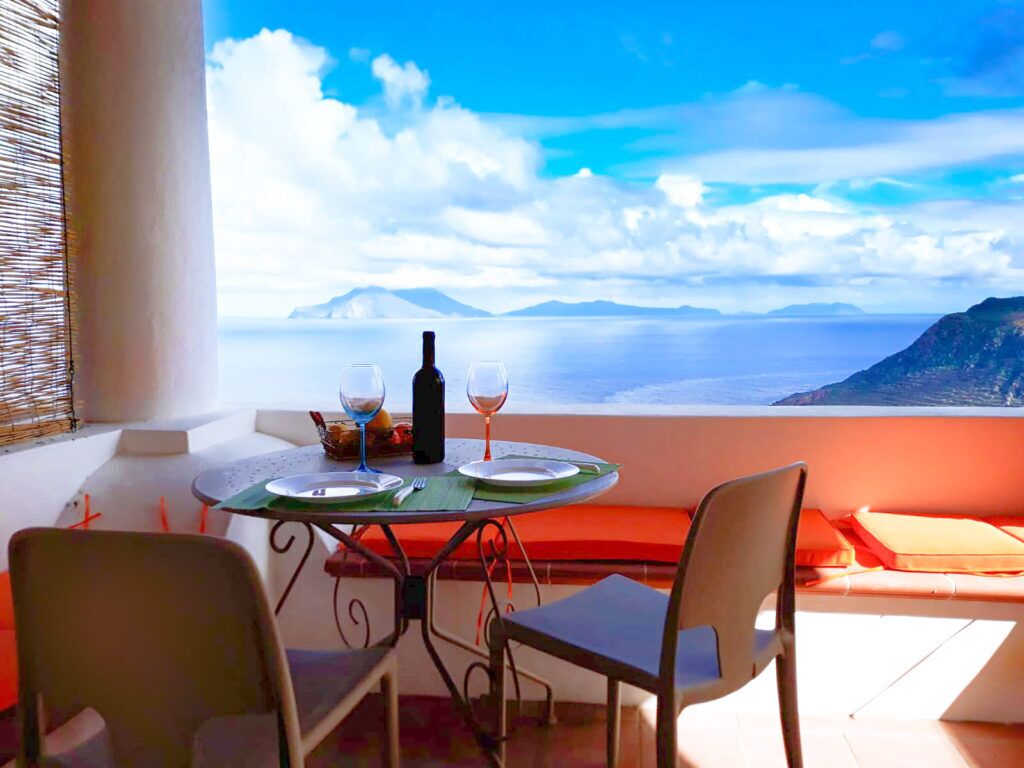 Une table dressée pour deux personnes, avec une bouteille et des verres à vin, offrant une vue panoramique sur la mer et les montagnes.