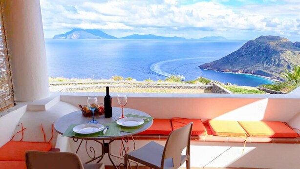 Un balcon avec une table dressée pour deux personnes, offrant une vue sur la côte, avec une montagne et la mer en toile de fond.
