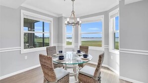 Un coin repas avec une table en verre et des chaises en osier, de grandes fenêtres donnant sur un champ, et un lustre.