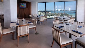 Un vaste espace de restauration avec de grandes tables et des chaises, un écran de télévision et de grandes fenêtres donnant sur un paysage urbain.