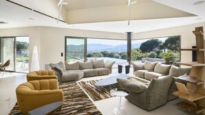 A spacious living room with a modern design, featuring a large sofa set, a coffee table, and a view of the outdoors through large windows.
