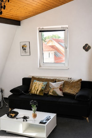 Una sala de estar con un sofá negro, una mesa de centro blanca, un cuadro enmarcado y una ventana con vista a un edificio.