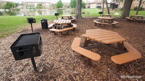 An outdoor picnic area with wooden benches and a grill, surrounded by trees and a body of water. - Aqua Sol is a fun family adventure! (Winter Garden)
