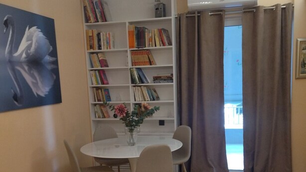 A modern living room with a sofa, a dining table, and a bookshelf. - Swan Paradise Glyfada Apartment  (Glyfada)