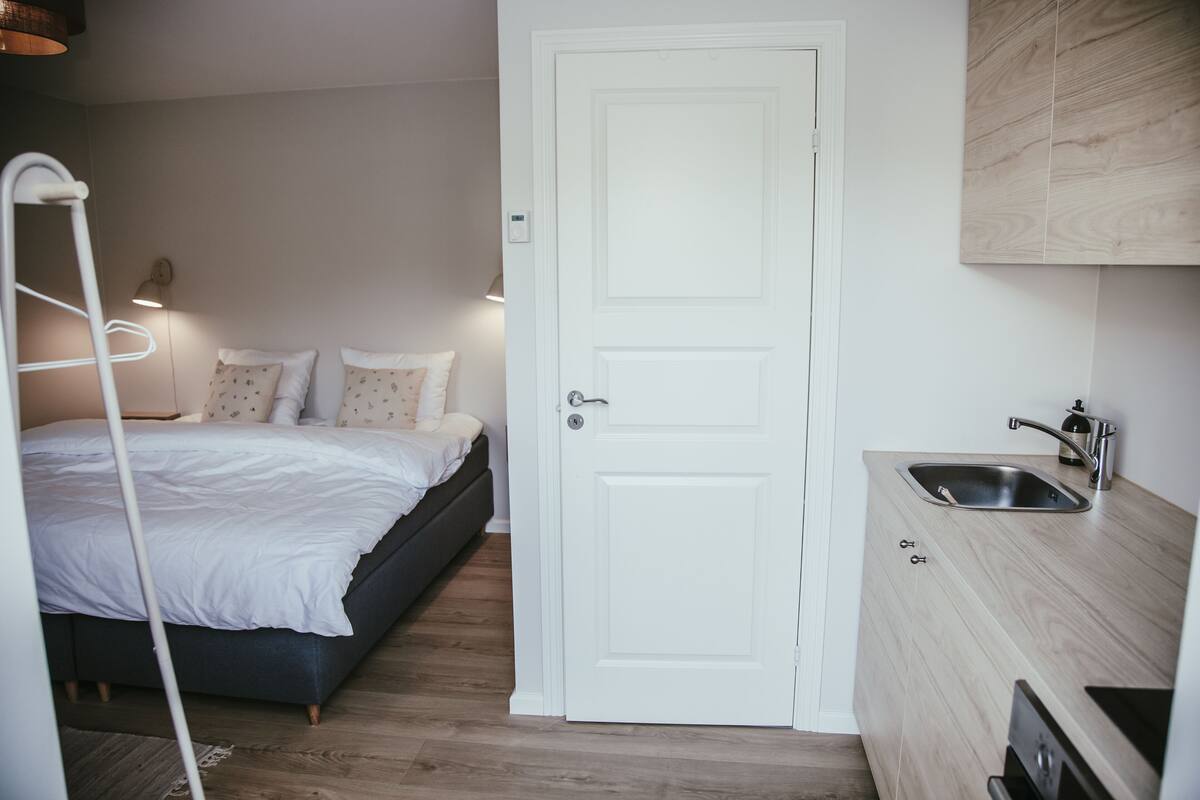A compact bedroom with a bed, bedside lamp, and a small kitchenette with a sink and cabinets.
