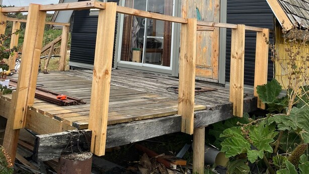 Une petite cabane aux couleurs sombres, dotée d’un toit à forte pente, d’une terrasse en bois et d’une cheminée d’où s’échappe de la fumée.