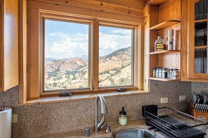 A kitchen with a large window, granite countertop, stainless steel sink, and wooden cabinets. - 4 Kings Log Cabin 150 Acres Spectacular Views (Three Rivers)