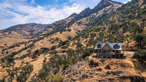 A cabin with a metal roof, surrounded by dry grass and sparse trees, with a mountain in the background. - 4 Kings Log Cabin 150 Acres Spectacular Views (Three Rivers)