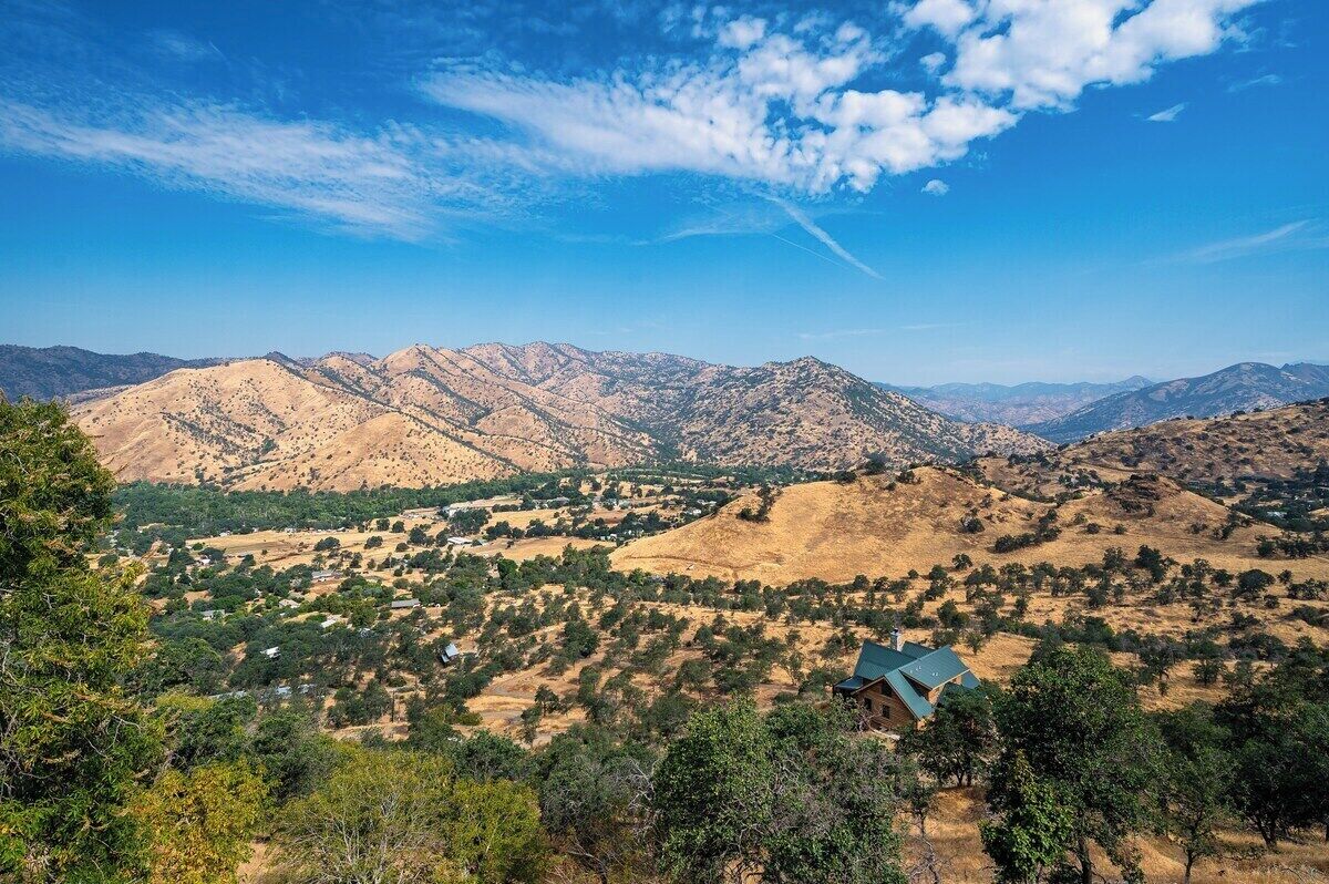 Un paisaje montañoso con un valle, una casa pequeña de techo azul y árboles dispersos.