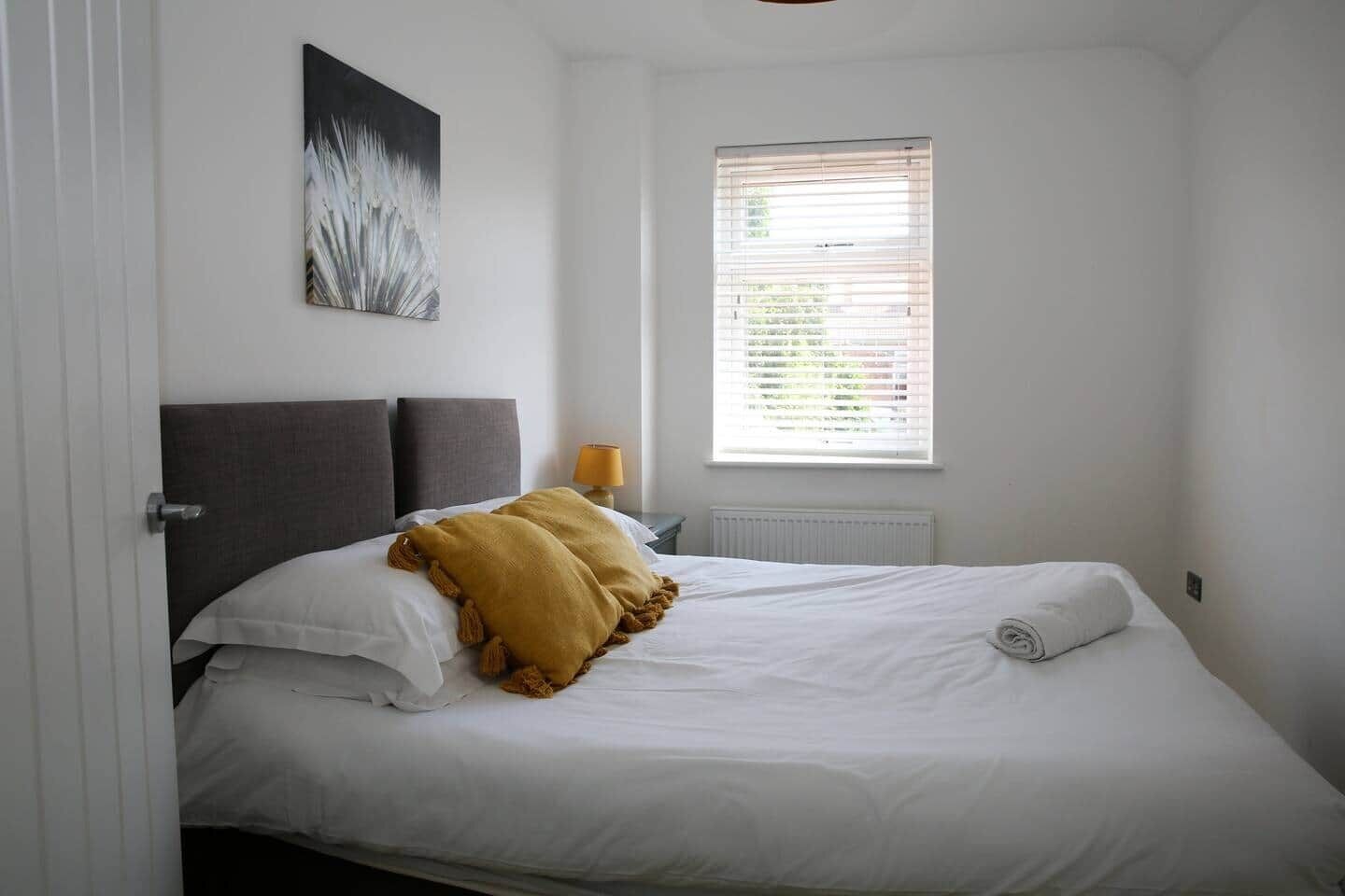 Un dormitorio con cama, ventana con persianas, lámpara y un cuadro en la pared.
