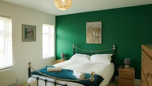 A bedroom with a green wall, a bed with white linens, a wooden nightstand, and a decorative lamp. - Myana Nest (Lowestoft)