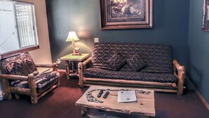 A cozy living room with a patterned sofa, a wooden armchair, and a small table with remote controls and a book. - Timber Creek Chalets: 6 1 Bedroom Chalet (Estes Park)