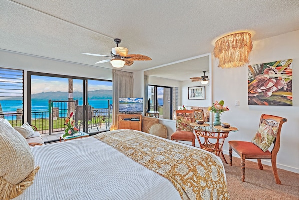 A spacious bedroom with a large bed, a ceiling fan, a television, and a balcony with a view of the ocean.