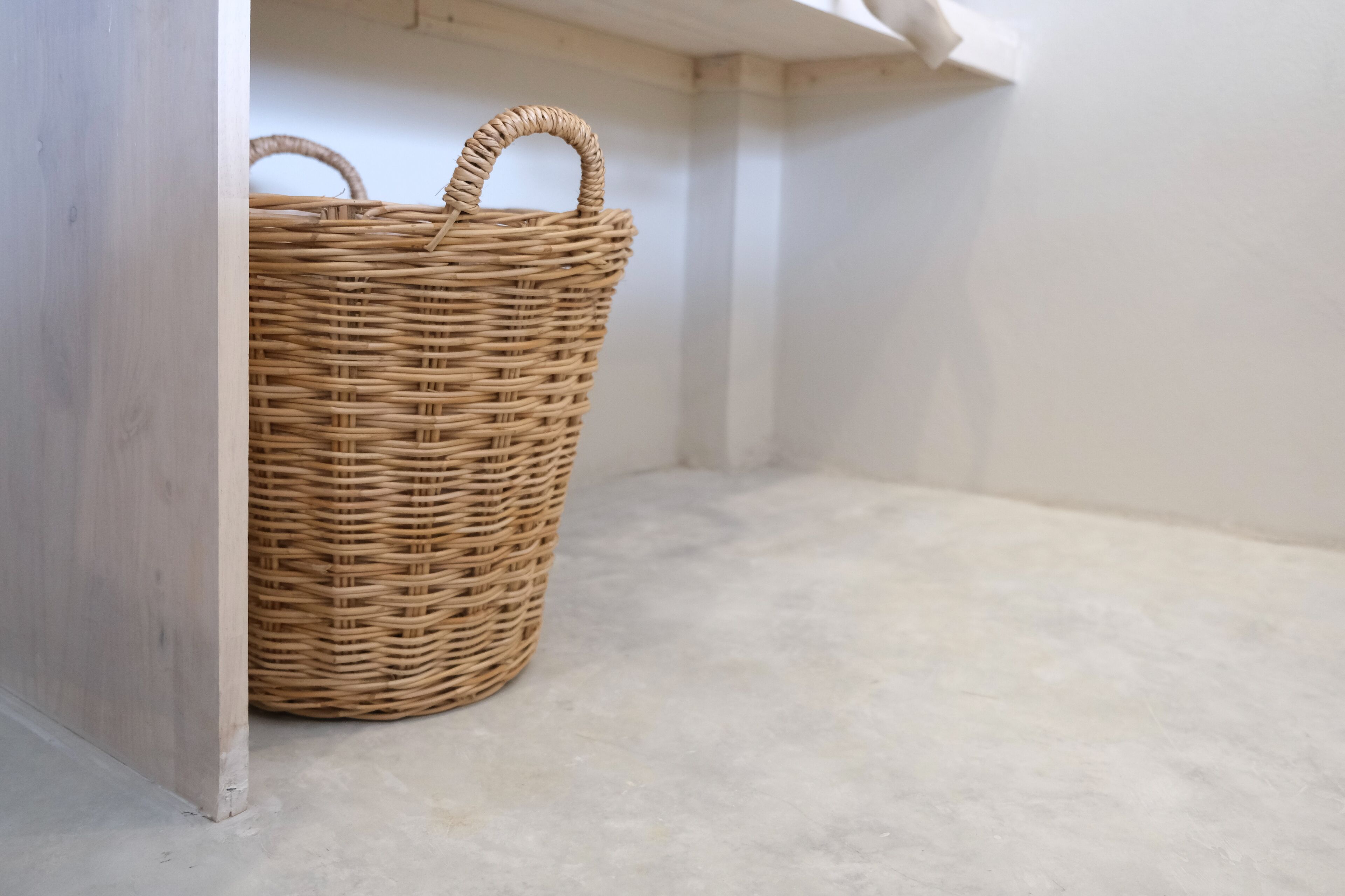 Un panier en osier, placé dans un coin d’une pièce dont le sol est simple et les murs sont blancs.