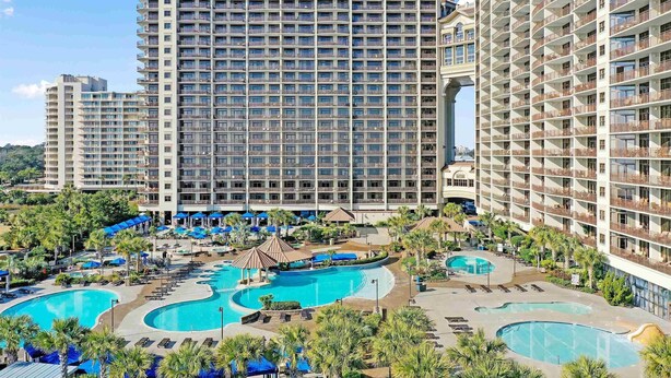 A hotel with multiple high-rise buildings, a central pool area with multiple pools, and surrounding greenery.