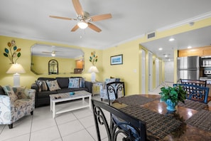 A living room with a sofa, coffee table, and dining table with chairs. There is a ceiling fan, a mirror, and a kitchen area with appliances.