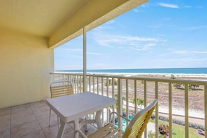 Een balkon met een witte tafel en stoelen, met uitzicht op een strand en een weg met begroeiing.