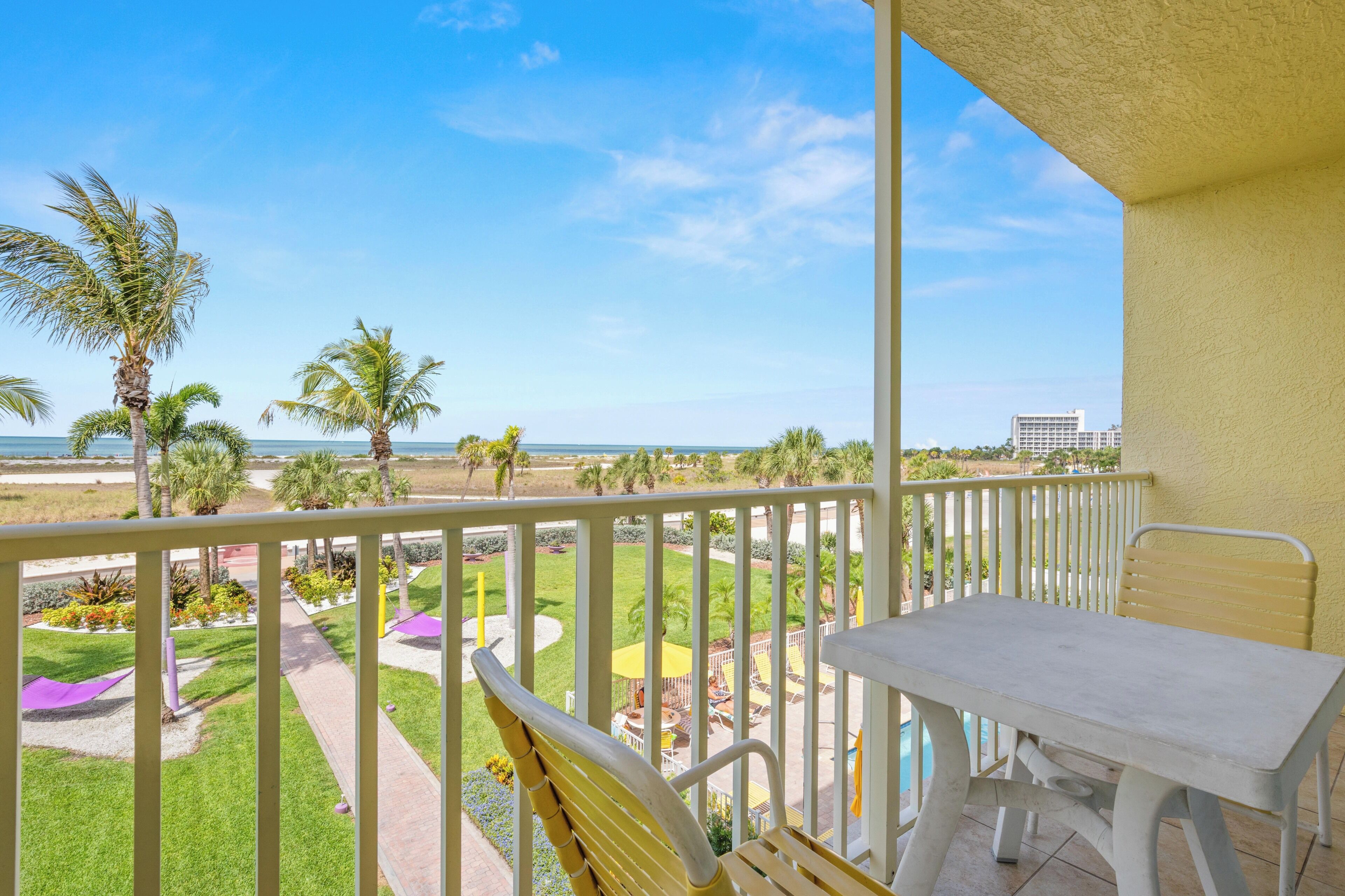 A balcony with a table and chair, overlooking a landscaped area with palm trees and a view of the ocean.