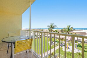 A balcony with a glass table and yellow chair, overlooking a beach and palm trees.