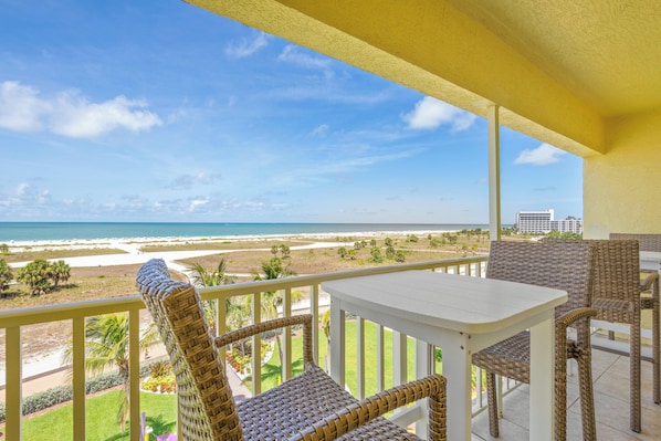 A balcony with wicker furniture, a white table, and a view of the beach and ocean.