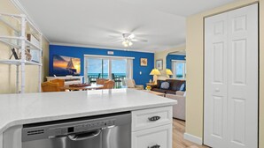 A kitchen with a dishwasher, a dining area with a table and chairs, a living room with a sofa and a lamp, and a view of the ocean through large windows.