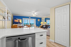 A kitchen with a dishwasher, a dining area with a table and chairs, a living room with a sofa and a lamp, and a view of the ocean through large windows.