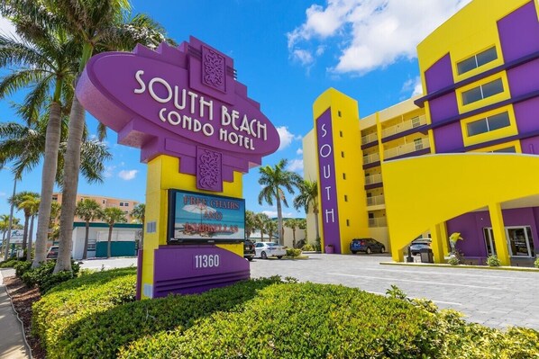 The South Beach Condo Hotel with a prominent purple and yellow sign, a parking area, and palm trees in the background. - 🌅 South Beach Condo #408  Spacious Gulf Front Escape for Families & Friends (Treasure Island)