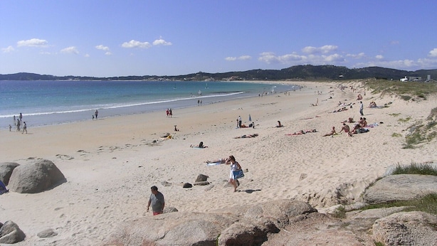 A wide sandy beach with people sunbathing and walking, large rocks scattered around, and a clear blue sky. - Apartamento EN EL Rural CON Terraza EN Sanxenxo, Cerca DEL MAR (Rebel- Vilalonga, Sanxenxo)