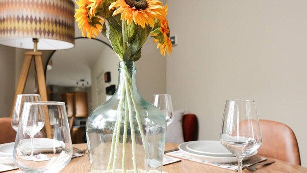 Un tavolo da pranzo con un vaso di vetro trasparente pieno di girasoli, una lampada e bicchieri da vino vuoti.
