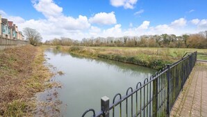 Un fiume tranquillo con una ringhiera metallica sulla destra, circondato da aree verdi e alberi sotto un cielo sereno.