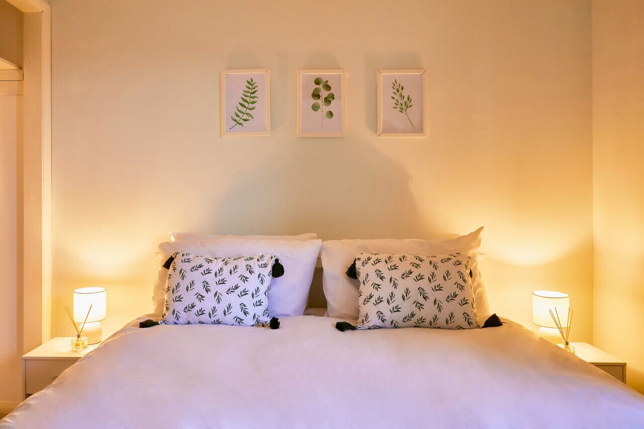 Un lit bien fait, avec une literie blanche et des coussins à motifs, est encadré de deux tables de chevet sur lesquelles reposent des lampes. Trois reproductions encadrées de gravures botaniques sont accrochées au mur au-dessus du lit.