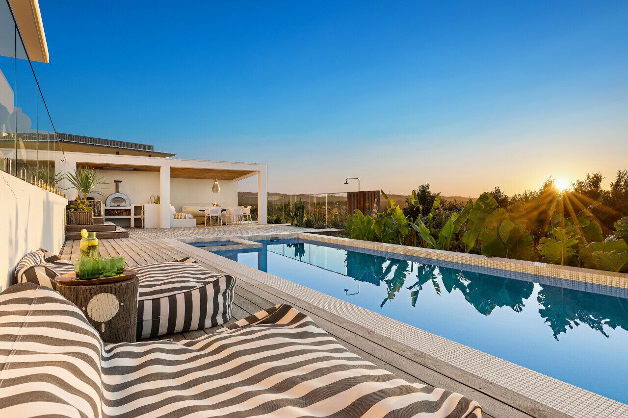 Un área de piscina moderna al aire libre con vistas despejadas al atardecer, un espacio de descanso con cojines a rayas y una zona de barbacoa.