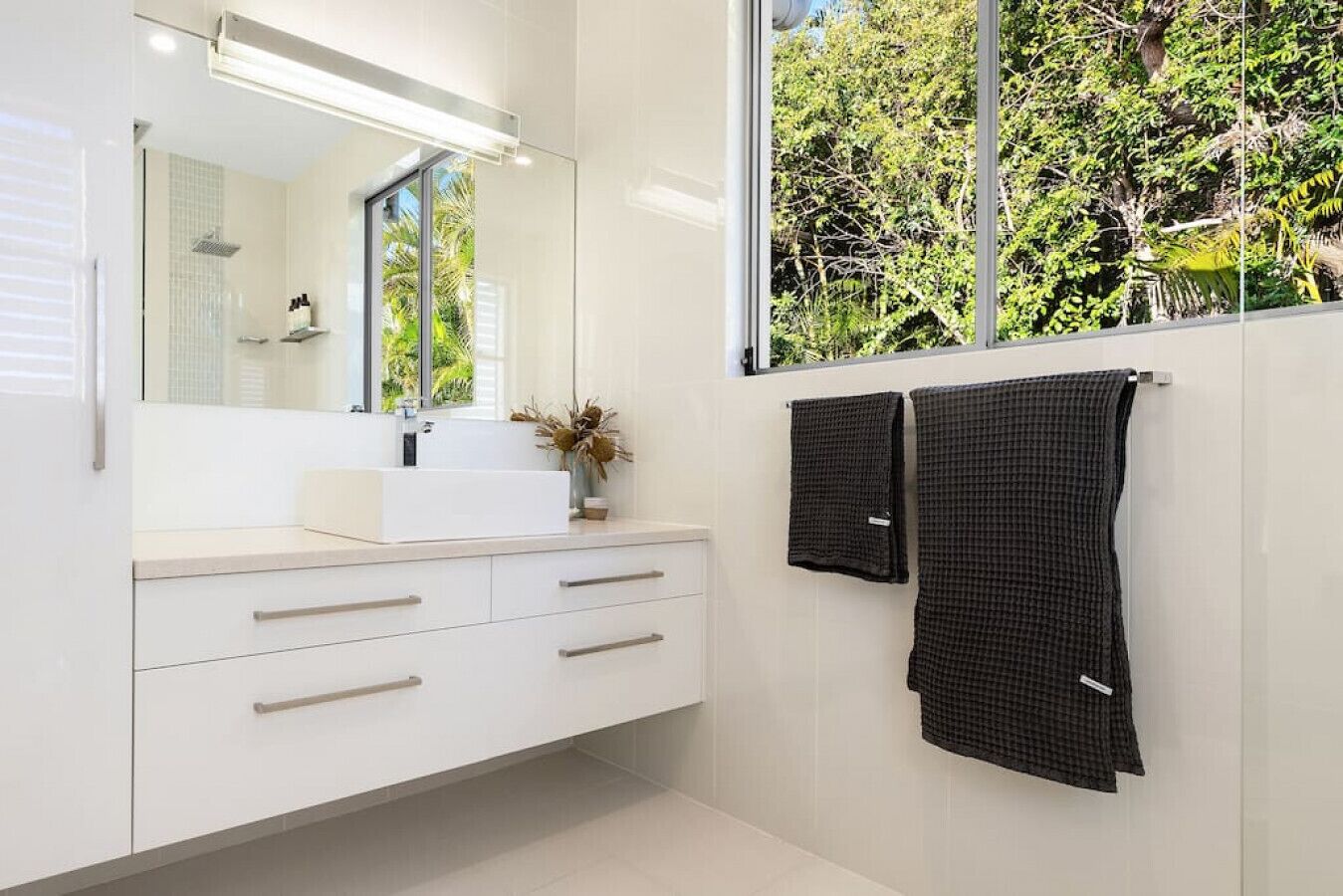 A modern bathroom with a large mirror, white cabinetry, and a window with a view of trees.
