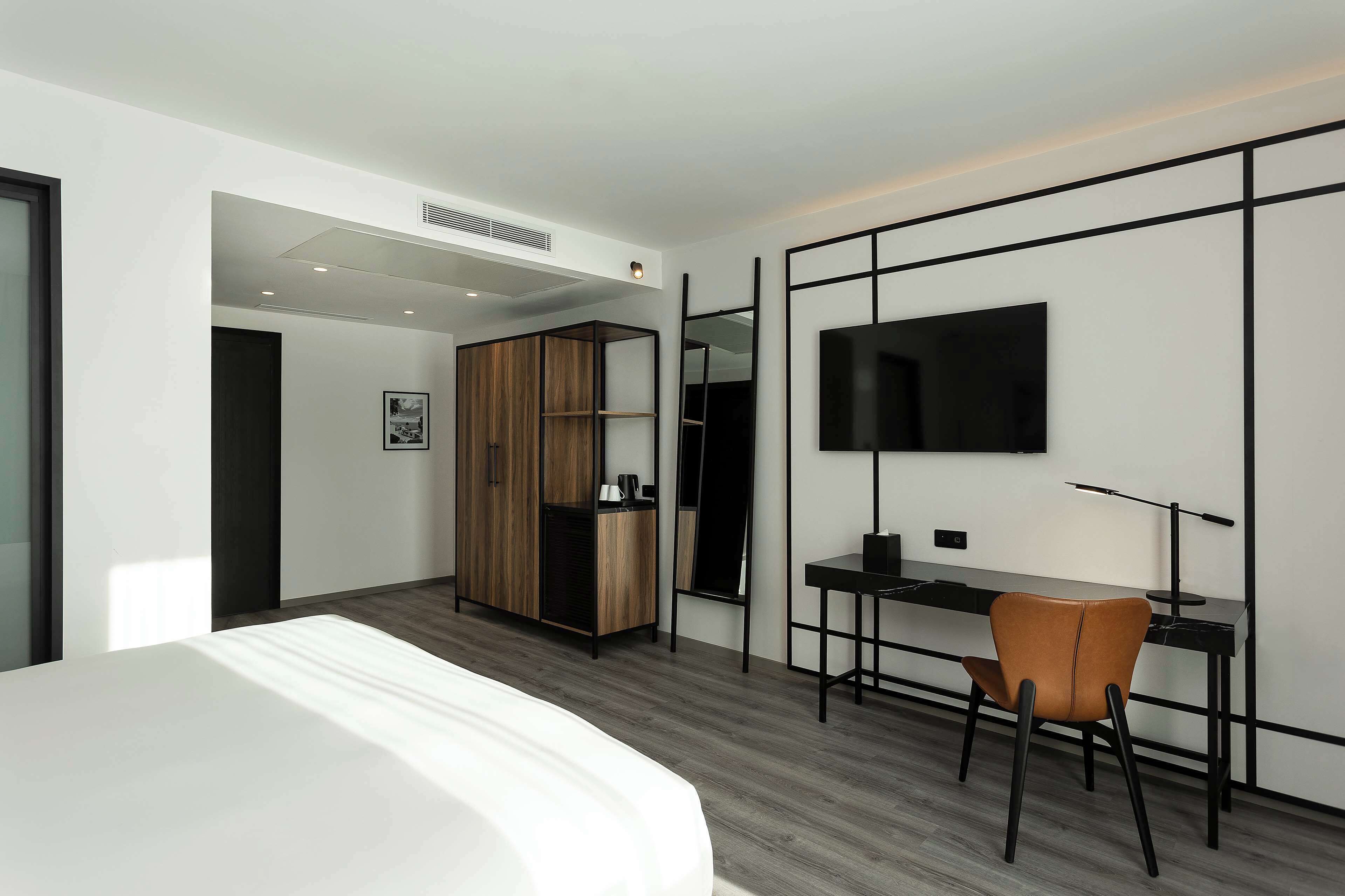 Quarto de hotel moderno com cama, escrivaninha, cadeira, guarda-roupa e televisão na parede.