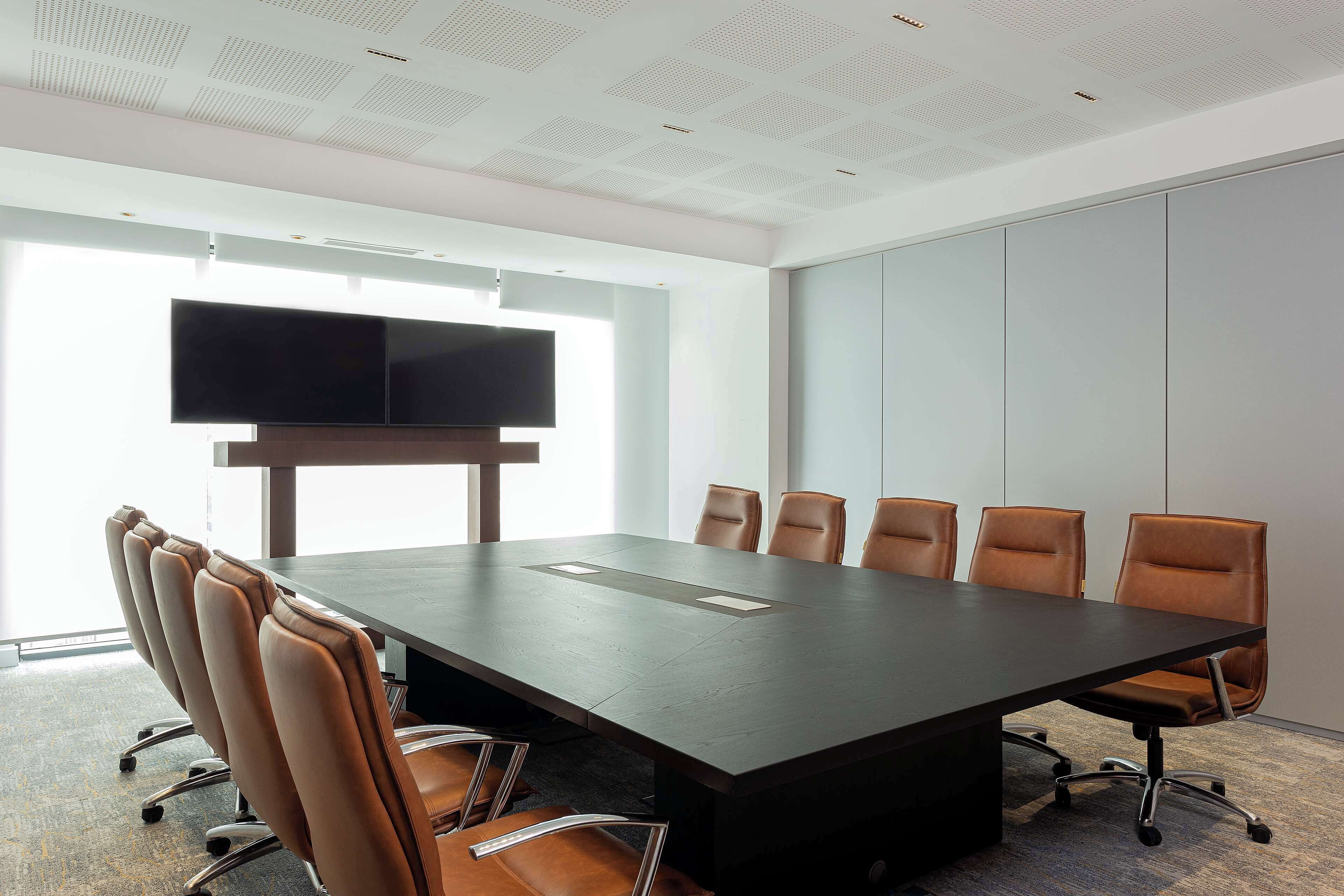 A conference room with a long table, six chairs, a flat-screen TV mounted on the wall, and large windows providing natural light.