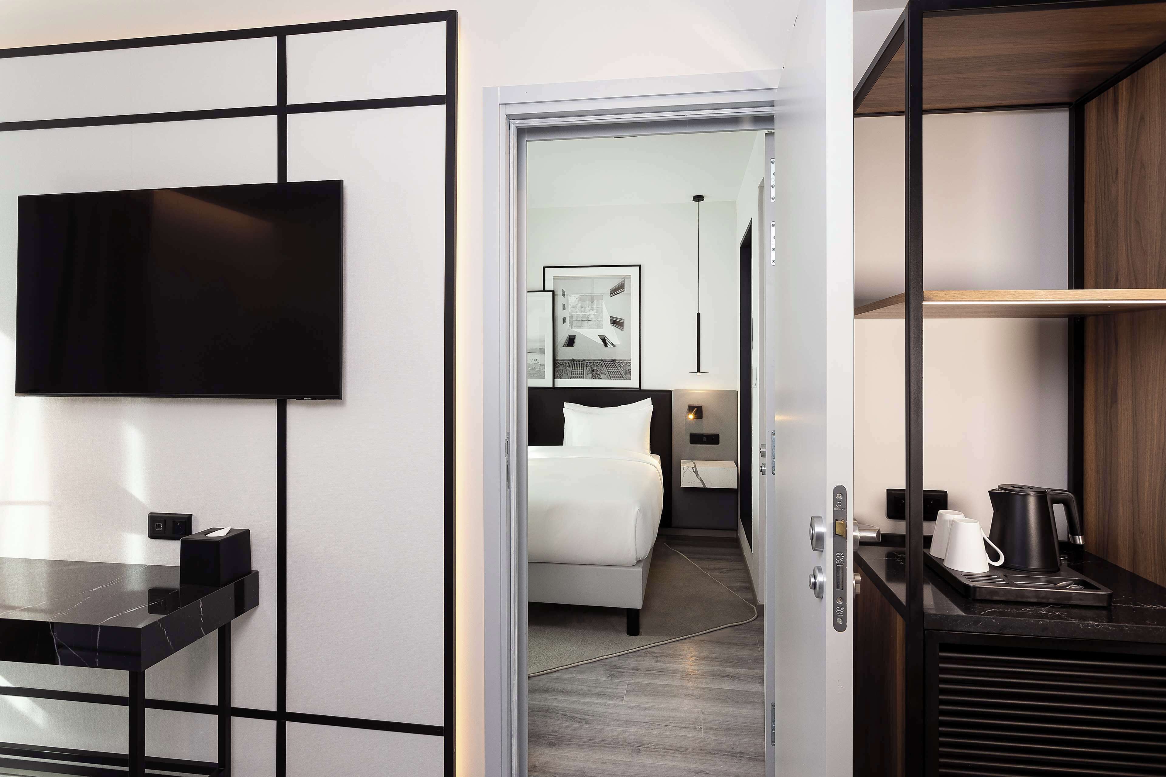 Une chambre d’hôtel moderne avec un téléviseur à écran noir, un lit avec une literie blanche, une table de chevet avec une bouilloire et une machine à café.