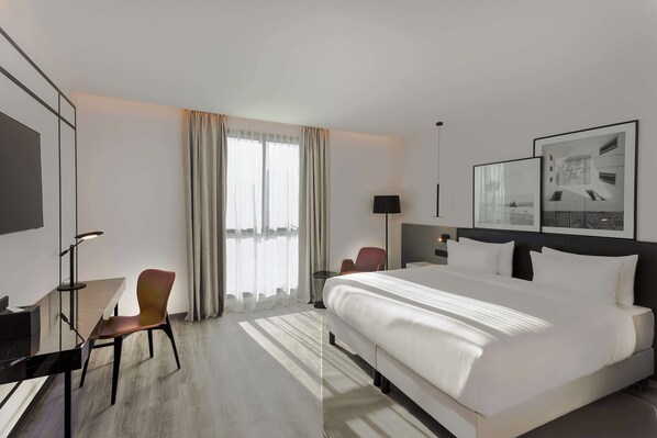 Habitación de hotel con una cama grande, un escritorio, una silla y un ventanal con cortinas.