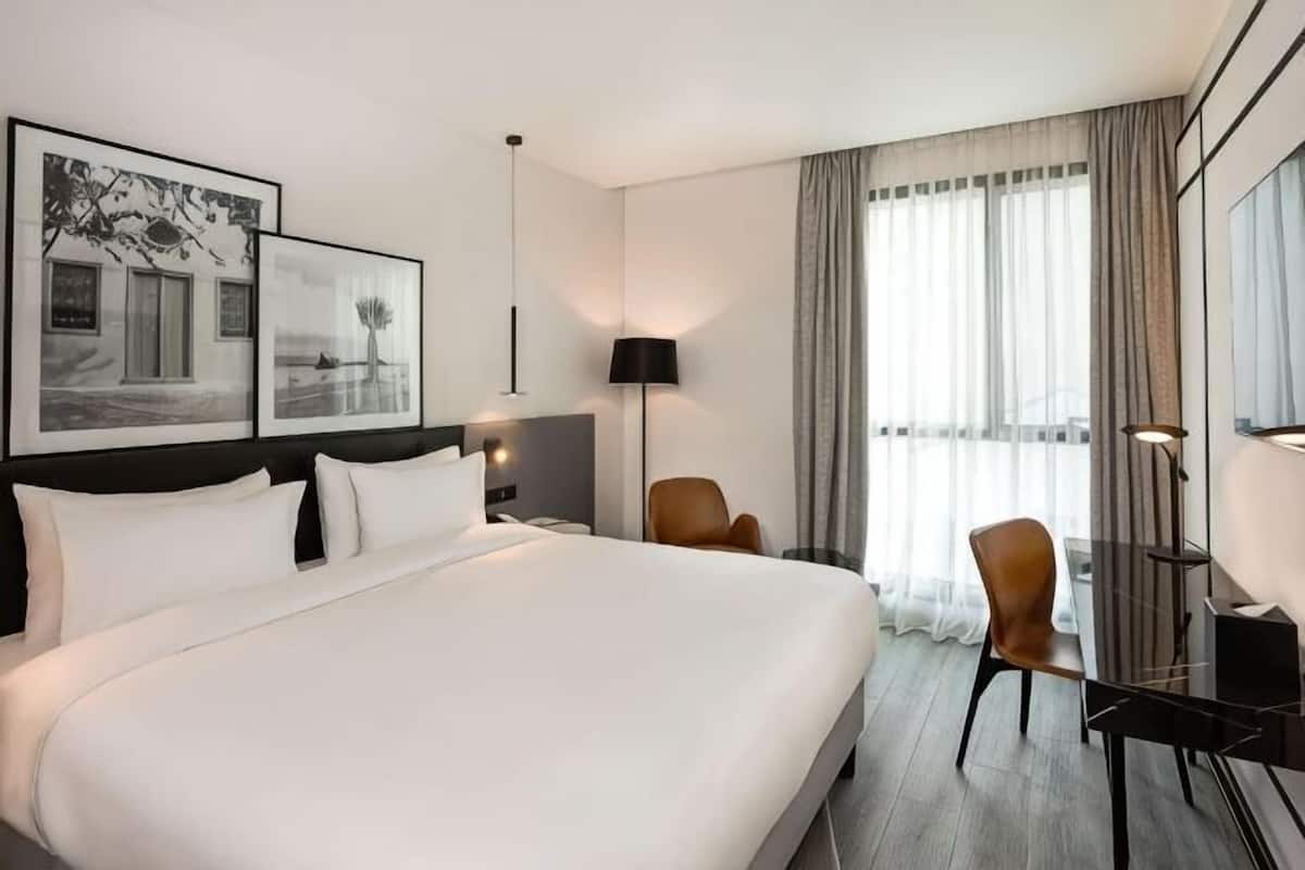 Ein Hotelzimmer mit einem großen Bett, einem Schreibtisch, zwei Stühlen, einer Lampe und gerahmten Bildern an der Wand.