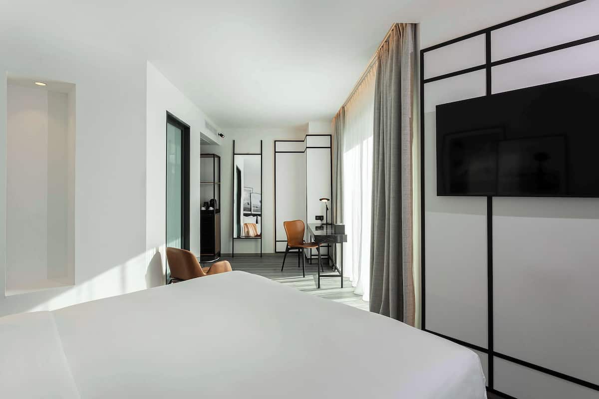 Ein modernes Hotelzimmer mit einem großen Bett, einem Schreibtisch, einem Sessel und einem an der Wand montierten Fernseher.