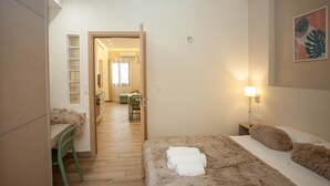 A hotel room with a bed, a desk, and a chair. There is a door leading to another room. - Blue Jasmine II (Chania)