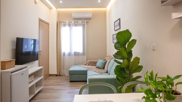 A modern living room with a flat-screen TV, a sofa, a dining table with a tray, and a large potted plant. - Blue Jasmine II (Chania)