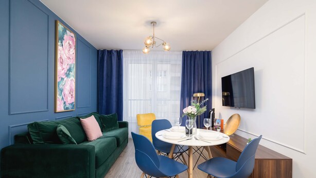 Un salon moderne avec un canapé vert, des fauteuils bleus, une table à manger dressée pour quatre personnes et une grande fenêtre ornée de rideaux bleus.