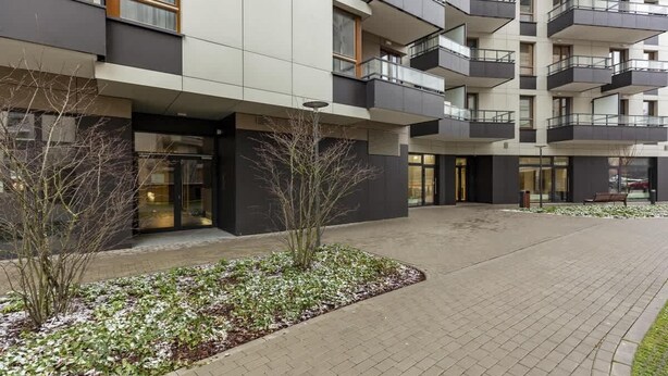 A modern multi-story building with balconies, a paved entrance area, and a small garden with plants and snow. - Stylish Apartment in the Center | Air Conditioning and Balcony (Warszawa)