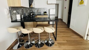 Eine moderne Küche mit schwarzem Arbeitsplatz, einer Kaffeemaschine und einem Kühlschrank. Drei Barhocker mit Chromfüßen ergänzen das Bild.