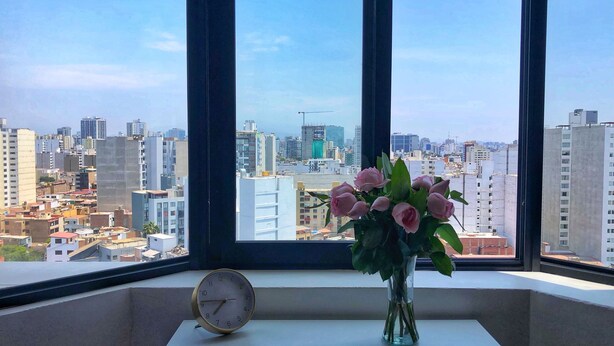 Ein Fenster mit Blick auf eine Stadtlandschaft, eine Vase mit Blumen und eine Uhr auf einem Tisch.