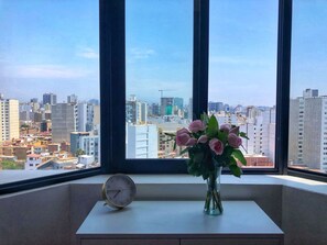 A window with a view of a cityscape, a vase with flowers, and a clock on a table. - Loft near the airport, in the best area of ​​Lima (Magdalena del Mar)